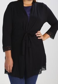 Cardigan nero con legatura frontale, maniche a tre quarti e bordo in pizzo ai polsini e all'orlo, realizzato in un tessuto morbido e leggero.