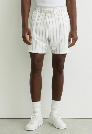 Homme portant un short blanc à fines rayures verticales, des chaussettes blanches et des baskets blanches, debout contre un mur blanc uni.