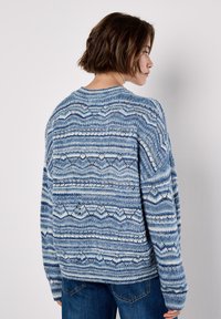 Woman with short brown hair wearing a blue patterned knit sweater and jeans, facing sideways against a plain white background.