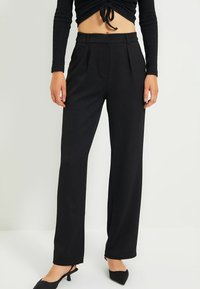 Pantalons larges noirs avec une coupe ajustée, confectionnés en tissu doux et ornés de plis à l'avant. Associés à des escarpins noirs à bout pointu.