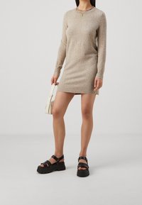Abito in maglia beige con maniche lunghe, polsini e orlo a costine. Abbinato a sandali neri chunky. La modella tiene una piccola borsa bianca.