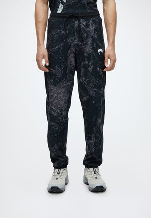 Hombre con pantalones jogger negros y grises con patrón, cintura con cordón y zapatos de senderismo gris claro sobre fondo blanco.