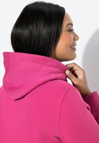 Bluza z kapturem w kolorze fuksji o regulowanym sznurku przy kapturze, ściągaczach na mankietach oraz miękkiej bawełnianej tkaninie, widziana z boku przy szyi i ramieniu.