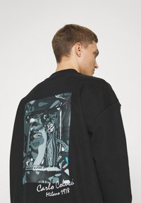 Zwarte sweatshirt met een grafische print op de achterkant met abstracte ontwerpen en tekst in het wit, die het merk en het jaar benadrukt.
