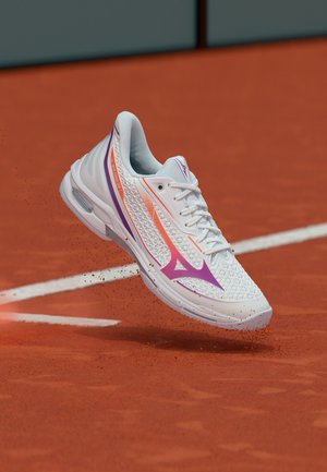 Chaussure de tennis blanche et violette soulevant de la poussière d'argile sur un court de tennis en terre battue rouge près d'une ligne blanche.