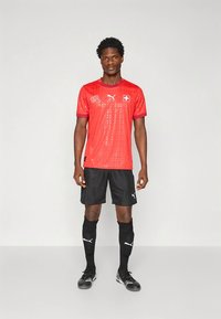 Piros rövid ujjú focimez texturált mintával, PUMA logóval és svájci zászló jelvénnyel. Fekete rövidnadrág, zokni és stoplis cipő egészíti ki az öltözéket.