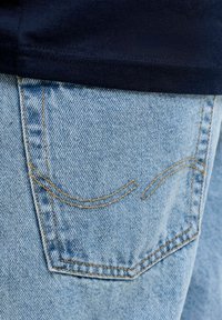 Poche arrière d'un jean en denim bleu clair avec des coutures orange et une partie d'une chemise bleu marine foncé visible au-dessus de la poche.
