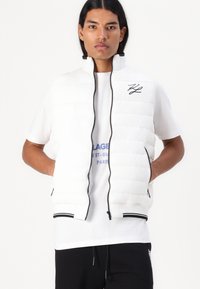 Balta paminkštinta liemenė su aukštu apykakle, juoda užtrauktuku ir kontrastingais juodais apvadu. Išsiuvinėtas logotipas ant krūtinės. Dėvima virš baltos marškinės.