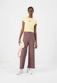 Kollane lühike T-särk tikandiga, kombineeritud laia jalgadega mauve värvi pükste ja valgete tossudega. Modell hoiab väikest kreemikat kotti.