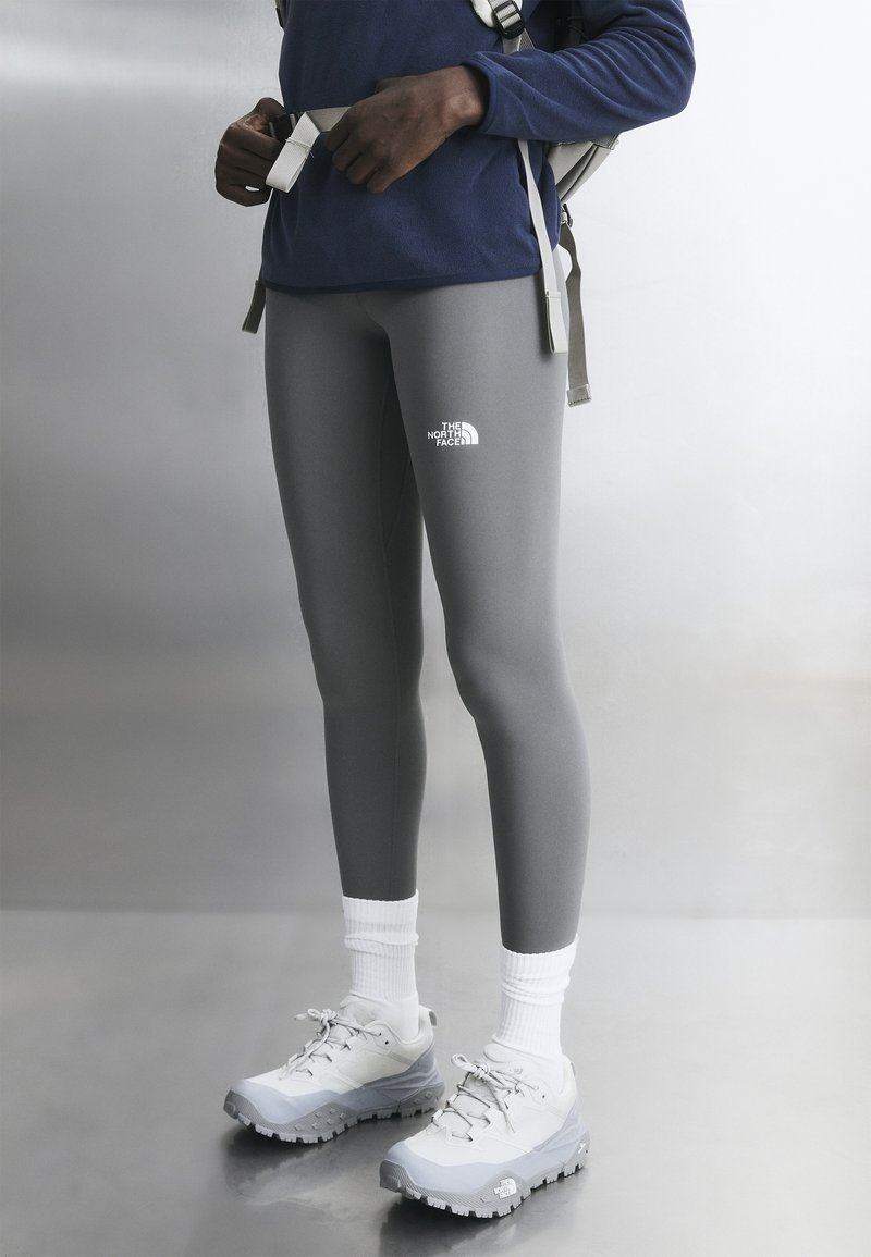 Henkilö, joka käyttää harmaita North Face -leggingssejä, valkoisia sukkia, valkoisia vaelluskenkiä ja säätää reput hihnoja yksiväristä taustaa vasten.