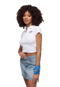 Polo shirt bianco a maniche corte con ricamo "NEWPORT BEACH", abbinato a una gonna di jeans leggera blu e una bandana stampata blu al polso.