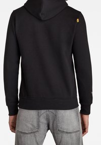 Sudadera negra con capucha, con dobladillo y puños acanalados. Presenta un pequeño logo amarillo en el hombro. El material parece suave y liso.