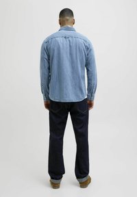 Camicia di denim azzurro chiaro con polsini abbottonati, abbinata a jeans blu scuro. La camicia presenta un orlo arrotondato e un colletto classico.
