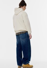 Beige hoodie med huva, lager på ett rutigt skjorta; lösa blå jeans med bakfickor; gula stövlar; brun stickad mössa.