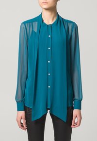 Blusa teal con maniche lunghe trasparenti, chiusura frontale con bottoni e colletto drappeggiato. Tessuto morbido con una consistenza liscia, con una leggera brillantezza.