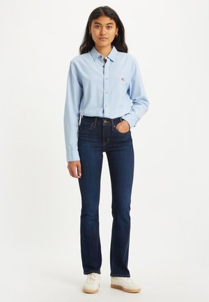 Chemise boutonnée bleu clair en coton, avec de longues manches et un petit logo. Portée avec un jean bleu foncé et des chaussures blanches.