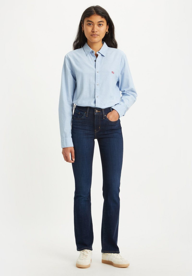 Chemise boutonnée bleu clair en coton, avec de longues manches et un petit logo. Portée avec un jean bleu foncé et des chaussures blanches.