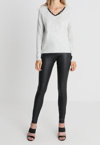 Helles graues Strickpullover mit V-Ausschnitt und schwarzem Spitzenbesatz, kombiniert mit schwarzen Leggings und schwarzen Riemchenschuhen. Glatte Textur und taillierte Passform.