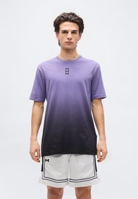 Νέος άνδρας που στέκεται μπροστά σε λευκό φόντο, φορώντας μπλούζα με διαβάθμιση μωβ και μαύρου και λευκά αθλητικά σορτς με μαύρη διακόσμηση.