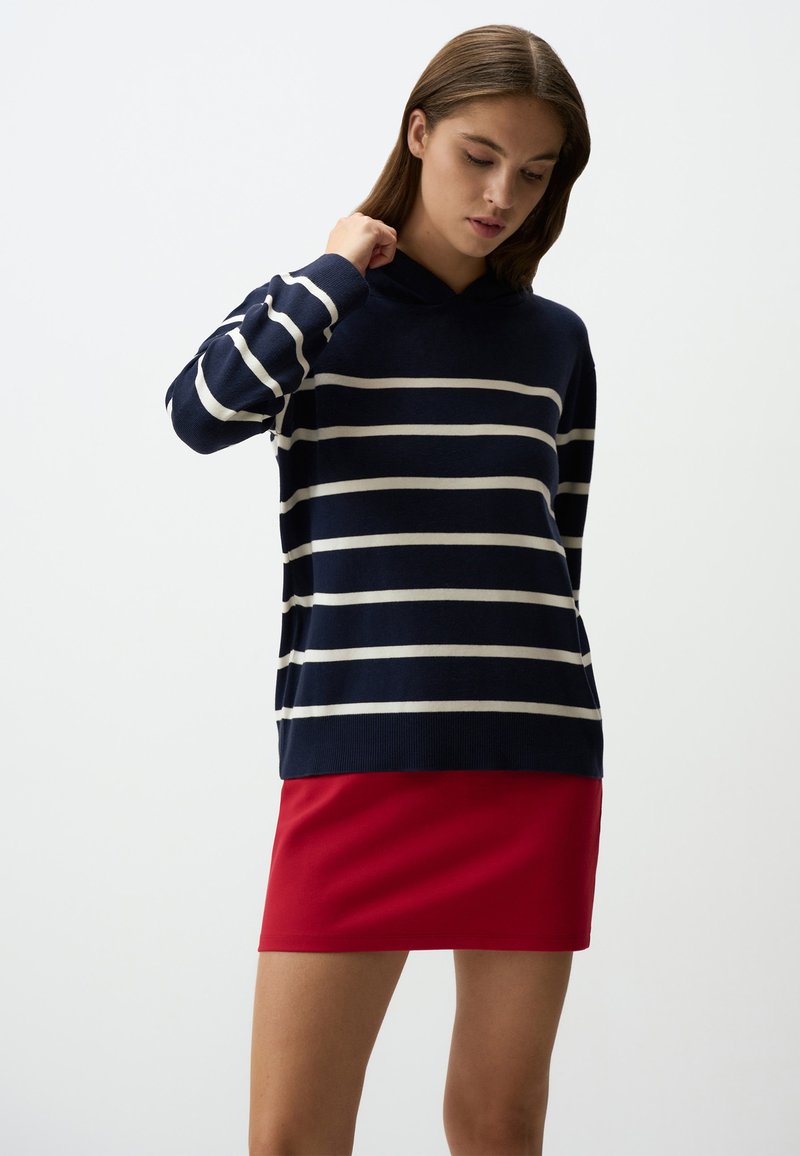 Maglione a righe blu navy con righe bianche e cappuccio, abbinato a una mini gonna rossa; tessuto morbido, vestibilità rilassata e maniche lunghe.