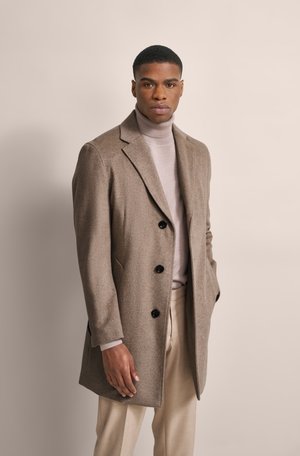 Brauner Wollmischungs-Mantel mit tailliertem Design, einreihiger Front, schwarzen Knöpfen und aufgeschlagenen Revers, getragen über einem hellen Rollkragenpullover.