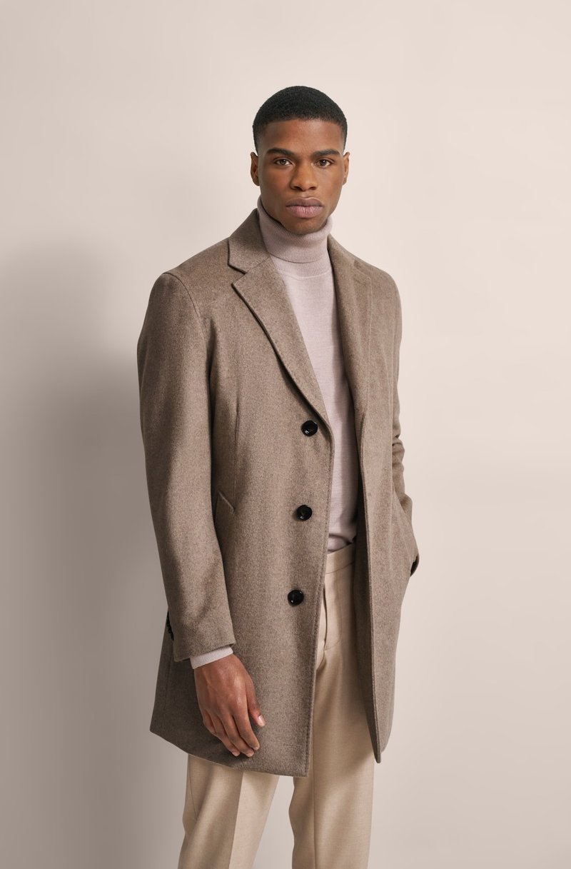 Cappotto a miscela di lana marrone con un design sartoriale, frontale monopetto, bottoni neri e rever a punta, indossato sopra un dolcevita chiaro.