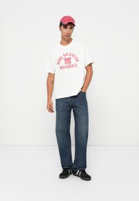 White oversized t-shirt with red text "Naval Air Station Pensacola, FL," paired with blue denim jeans and black sneakers. Red cap worn.