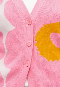 Rosa strikket cardigan med tre rosa knapper og store gule og hvite blomsterpatroner på stoffet.