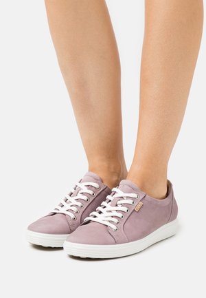 Jambes portant des baskets à lacets violet clair avec des semelles blanches et des lacets blancs, debout sur une surface blanche unie.