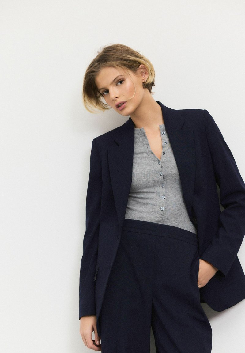 Navy blazer over a grey henley top with buttons, paired with navy trousers. Smooth fabric, tailored design, and a fitted silhouette.