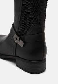 Bota de cuero negro con un panel de malla texturizada, una correa delgada en el tobillo con una hebilla circular y un tacón apilado para estabilidad y estilo.