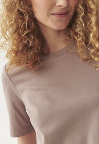 Světle hnědé tričko s krátkým rukávem vyrobené z měkké tkaniny, s klasickým kulatým výstřihem a hladkým povrchem. Bez viditelných vzorů nebo kovových doplňků.