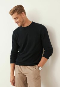 Pull noir à col rond en tricot doux, porté avec un pantalon beige clair. Une montre en argent accentue la tenue. Fond clair.