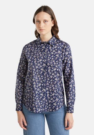 Camisa de manga larga azul marino con un estampado floral en colores claros, cierre de botones en la parte frontal y una textura suave; diseño de bajo curvado y cuello.