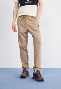 Beige jogger kalhoty s elastickým pasem a manžetami, s logem na boku. Nositeli spolu s tmavými teniskami na modrém povrchu.