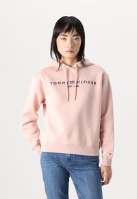 Sweat-shirt à capuche rose clair avec un ourlet et des poignets côtelés. Présente le logo "TOMMY HILFIGER" en noir à l'avant. Le matériau semble doux.