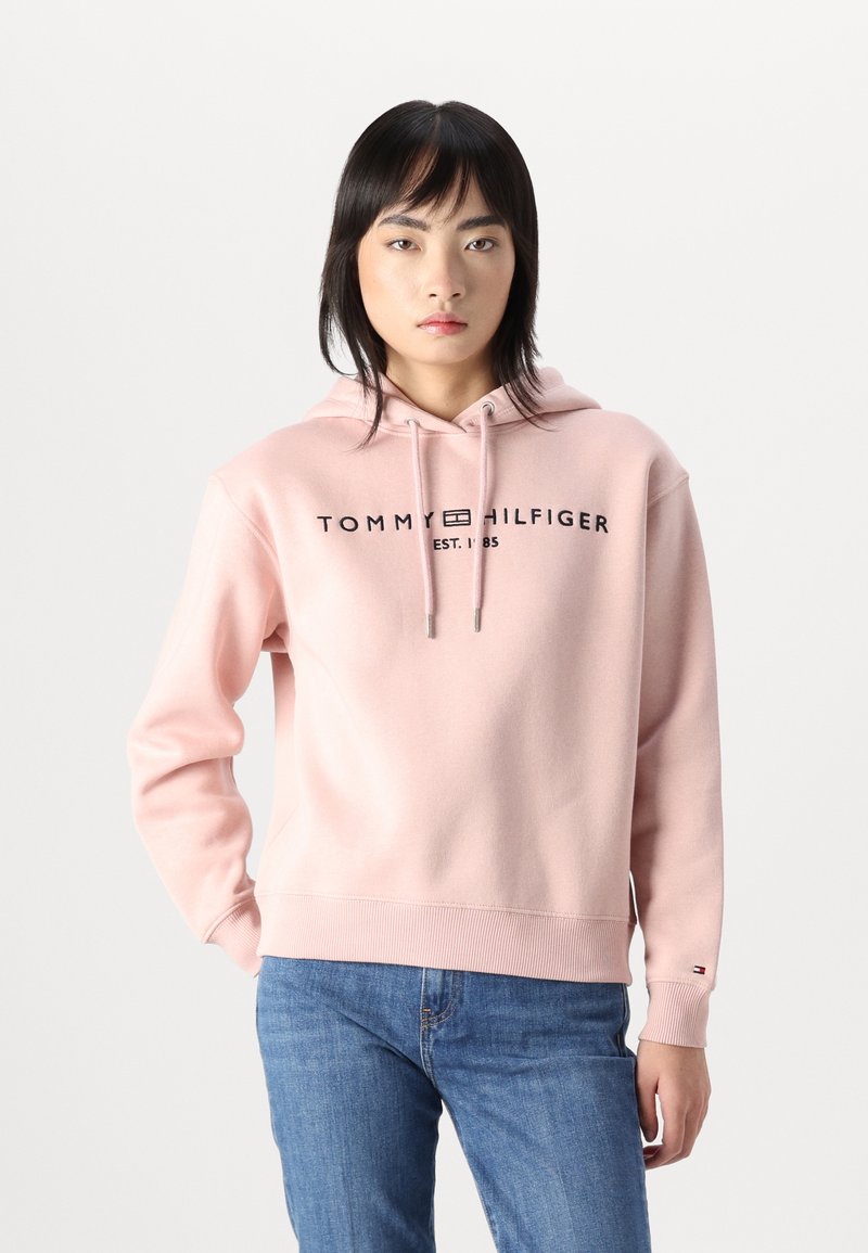 Sweat-shirt à capuche rose clair avec un ourlet et des poignets côtelés. Présente le logo "TOMMY HILFIGER" en noir à l'avant. Le matériau semble doux.
