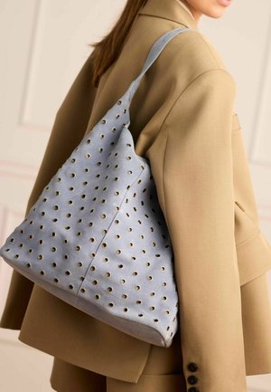 Donna con blazer color beige che porta una borsa a tracolla in suede azzurro chiaro con dettagli ad occhiello dorati.