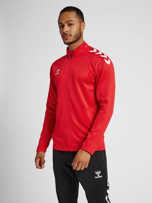 Homme portant une veste de sport rouge zippée avec des bandes chevrons blanches sur les épaules et un pantalon de sport noir avec un logo blanc sur la cuisse.