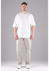 Weißes, übergroßes T-Shirt mit kurzen Ärmeln, kombiniert mit beigen Chinos und weißen Sneakern, vor einem neutralen grauen Hintergrund.