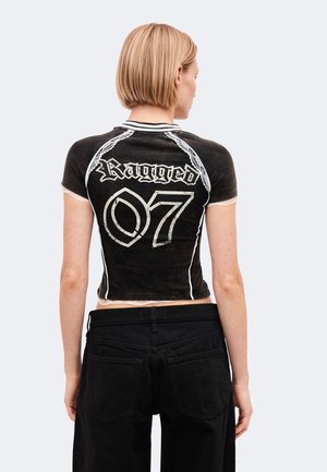 Personne aux cheveux blonds courts portant un t-shirt court noir avec "Ragged 07" dans le dos et un pantalon noir, tournée dos à la caméra.