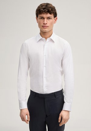 Młody mężczyzna o krótkich brązowych włosach, ubrany w białą, długorękawową koszulę z guzikami i czarne spodnie, stoi na jednolitym tle.