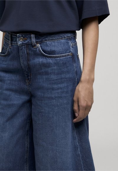Personne portant un jean large en denim bleu foncé et un t-shirt ample bleu foncé, avec un bras détendu le long du corps.
