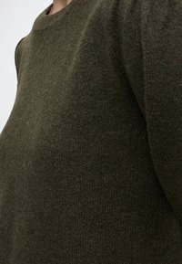 Gros plan d'une personne portant un pull en maille épais vert olive foncé avec un col rond haut.