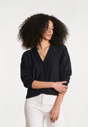 Femme aux cheveux bouclés portant un cardigan noir boutonné et un pantalon blanc, regardant vers sa gauche, les bras croisés à la taille.