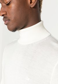Cremefarvet turtleneck sweater lavet af blødt strikket stof, med ribbet detalje ved kraven og et diskret logo på forsiden.