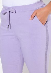 Ljust lila joggerbyxor med dragsko i midjan, sidofickor och en dekorativ silverkant längs sömmen. Mjuk tygtextur.
