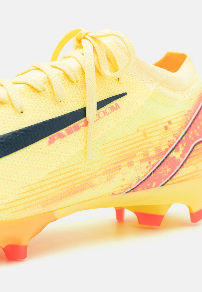 Geel voetbalschoen met een textuurmateriaal, zwarte accentstreep en levendige rood-oranje buitenzool met een dynamisch patroon. Vetersluiting.