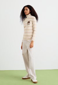 Haut à col roulé crème à manches longues avec des rayures horizontales marron et un logo de palmier, associé à un pantalon large beige clair et des chaussures blanches.