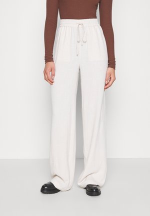 Person som bär lösa vita byxor med dragsko, brun långärmad topp och svarta klumpiga skor, stående mot en enkel bakgrund.